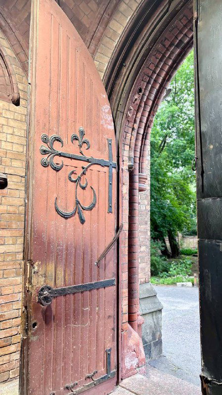 Открытая часть деревянной двери церковного портала с видом на зелёную аллею позади здания и часть кирпичной кладки у входа.