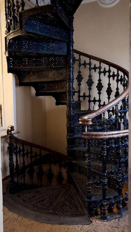Чёрная винтовая чугунная лестница с декоративными балясинами, ажурными элементами и тёмным деревянным поручнем в историческом интерьере усадьбы.