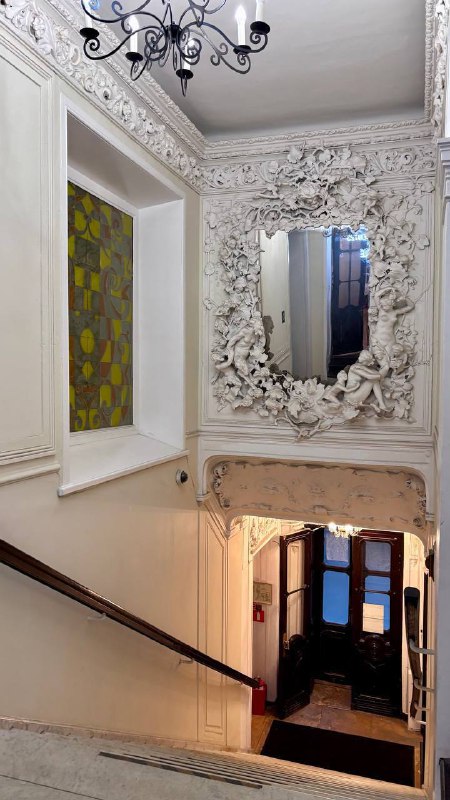 Ракурс лестничного холла усадьбы: большое резное зеркало, витражное окно, классическая люстра и декоративные орнаменты в одном кадре.