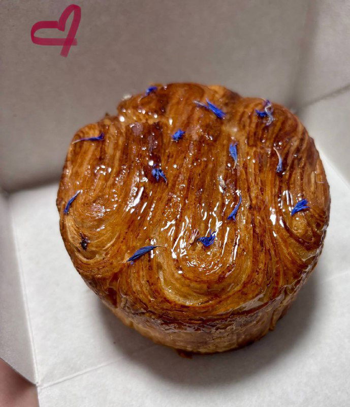 Круглая булочка с завихрениями теста сверху и синими съедобными цветами, вид сверху в коробке, текстура слоёного теста отчётлива.