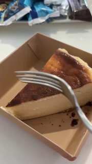 Кусок чизкейка Сан Себастьян в картонной коробочке, вилка рядом, плотная кремовая структура и поджаренная корочка сверху.