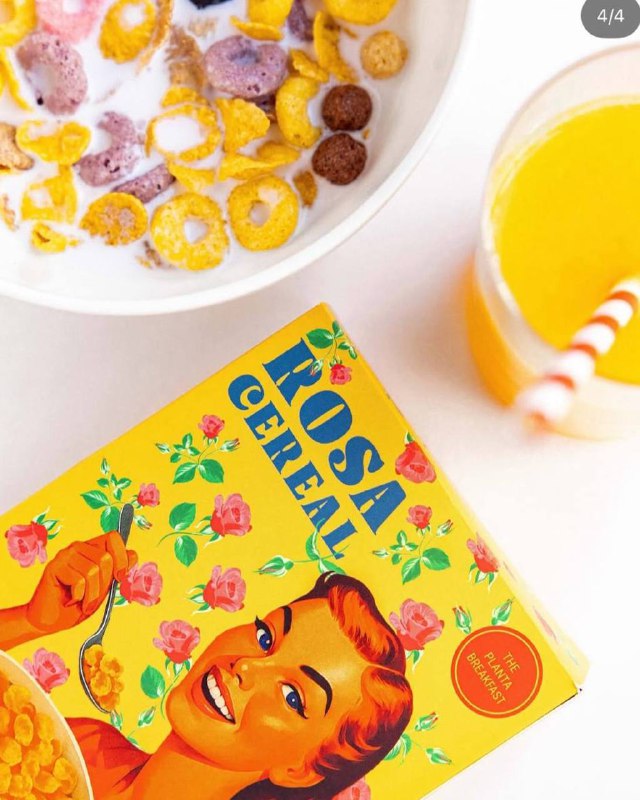 Крупный план хлопьев в миске и коробка фирменных Rosa Cereal на фоне апельсинового сока — стилизованная под американский завтрак композиция.