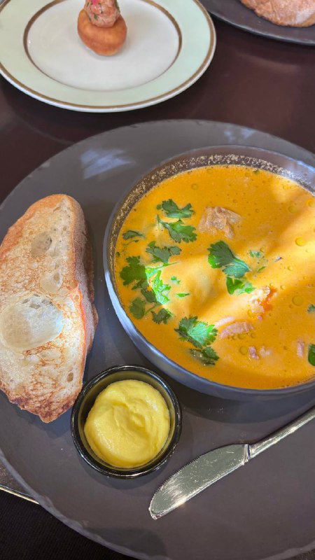Тарелка с ухой в Geraldine: кремовый оранжевый суп, зелень, кусочки хлеба и небольшая ёмкость с соусом — подача блюда на темной тарелке.