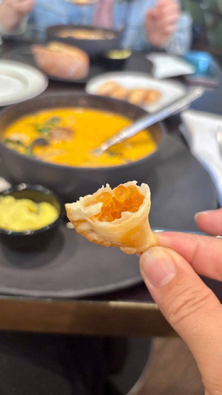 Кусочек чебурека в руках на фоне супа: видна текстура начинки, золотистая корочка и контекст дегустации за столом ресторана.