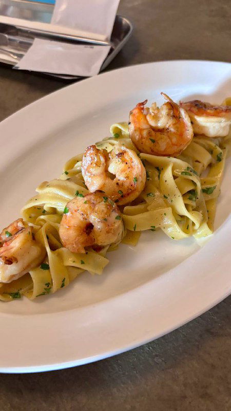 Крупный план фетучини с креветками на белой тарелке: видны сочные креветки, лента пасты и соус, подача на столе.