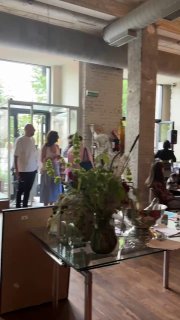 Другой ракурс зала бистро: у входа стоят люди, стеклянные столы и цветочные композиции на фоне кирпичных стен.