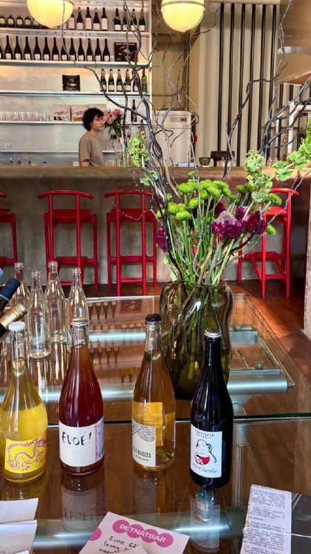 Барная стойка и витрина с бутылками лимонадов: стеклянные бутылки, букеты цветов и часть красного барного стула на переднем плане.