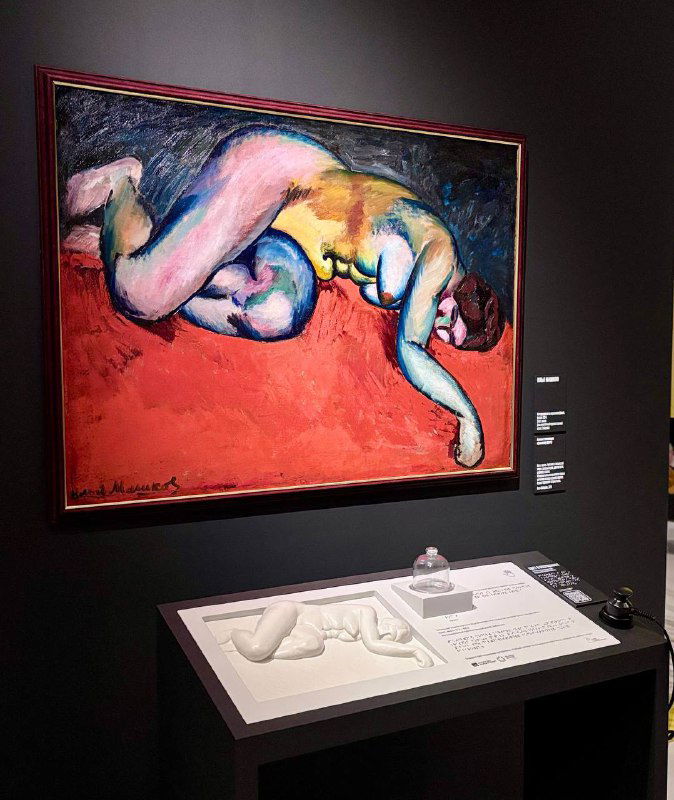 Большая картина с лежащей фигурой в насыщенных контрастных цветах, рядом визитка экспоната и музейная витрина с информацией.