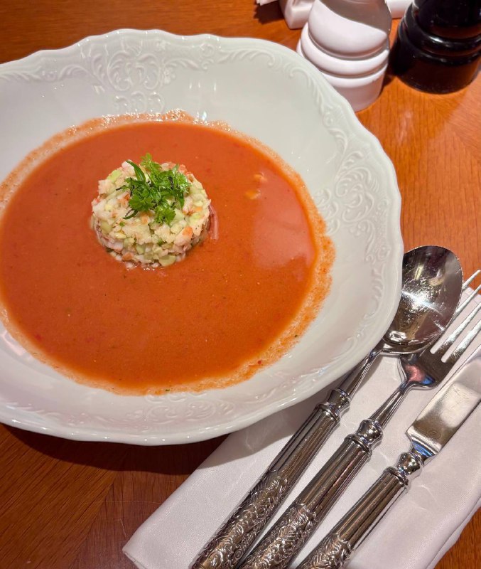 Тарелка гаспачо с центральным шариком гарнира, рядом сложенные серебряные столовые приборы и салфетка на деревянном столе ресторана.