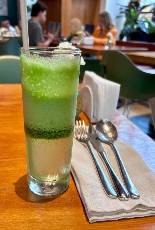 Высокий прозрачный бокал лимонада щавель-алоэ с заметным большим куском льда, на фоне приборов и интерьера кафе Flaner.