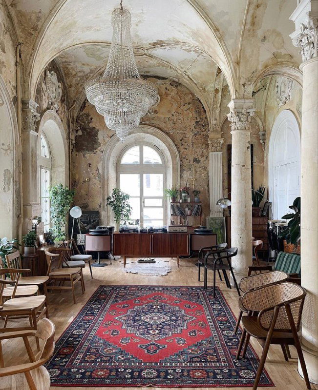 Большой зал с высокими арками, хрустальной люстрой и старинным ковром на полу, ощущение запущенной, но красивой архитектуры и света.