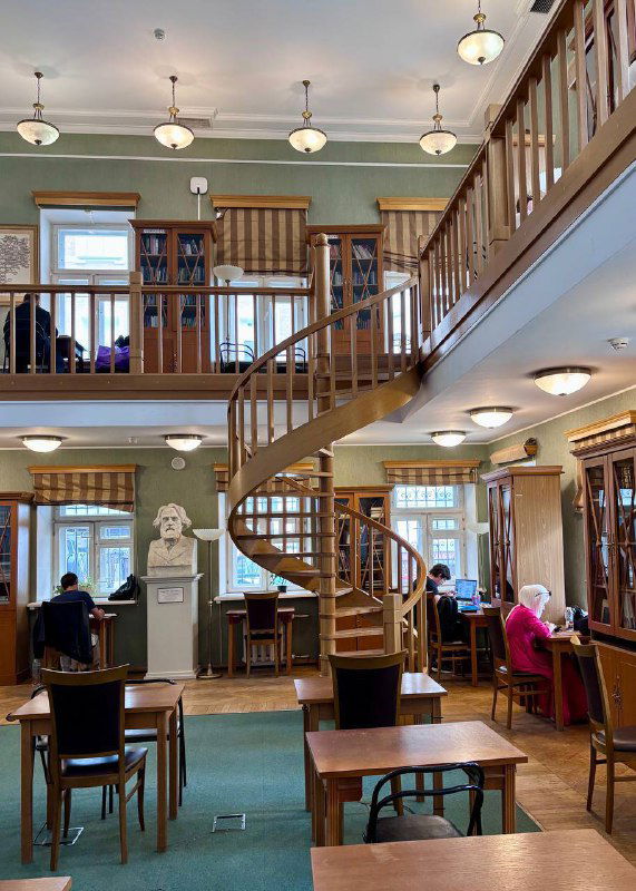 Читальный зал библиотеки им. Тургенева: столы, стеллажи, посетители и характерная спиральная лестница, вид на второй ярус пространства.