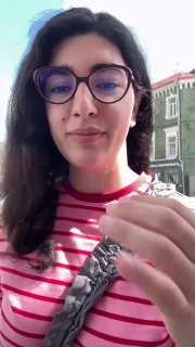 Селфи автора у окна кафе: она демонстрирует жест, на заднем плане видны фасады соседних зданий и дневной свет.