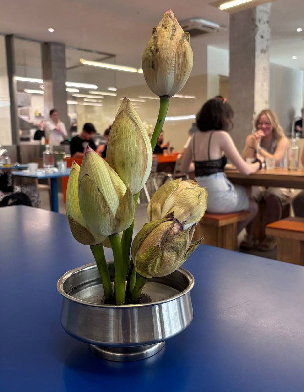 Небольшая композиция с букетом бутонов на столе кафе; видно пространство зала и посетителей на заднем плане в интерьере Zoe.