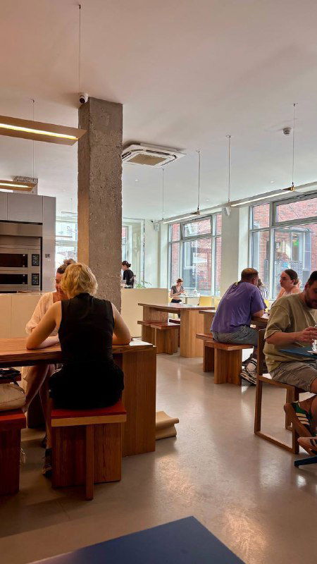 Интерьер пекарни Zoe: деревянные столы и стулья, большие окна и дневной свет, посетители за столиками — чистый современный стиль.