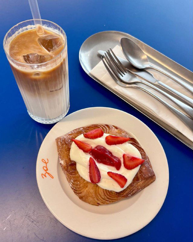 Латте айс и порция даниша с кремом и ломтиками клубники на белой тарелке — завтрачная сервировка на синем столе Zoe.