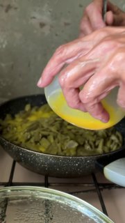 Руки вливают взбитые яйца в сковороду с жареной фасолью; видно процесс приготовления дзвов лоби на газовой плите во дворе.