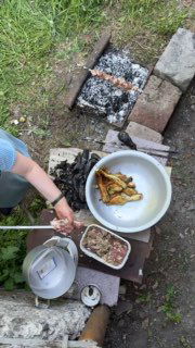 Общий план мангала с посудой и тарелками, на столе тарелка с запечёнными баклажанами и подготовленный фарш — рабочий процесс на улице.