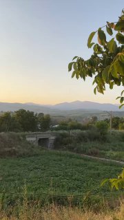 Панорамный вид на сельскую долину с полями, мостом и дальними холмами на закате: тихая летняя сельская сцена с мягким светом.