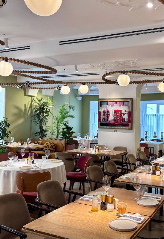 Интерьер ресторана Mouflon: светлый зал с круглыми лампами, зелёными акцентами, расставленными столами и сервировкой, уютная обстановка.