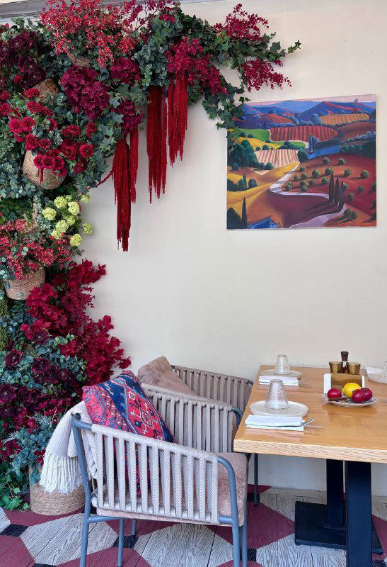 Декор ресторана: яркая цветочная инсталляция у стены и картина, рядом столик с простыми тарелками — уютный уголок для гостей.