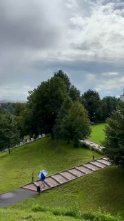 Тропинка в липовом парке усадьбы Дубровицы: ступени, зеленые деревья и человек с зонтом, отражение небольших луж на дорожке.