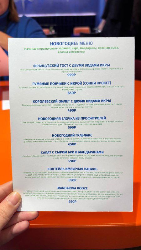 Рука держит распечатанное новогоднее меню кафе Скворец с перечнем праздничных блюд и цен — листовка сезона на столе