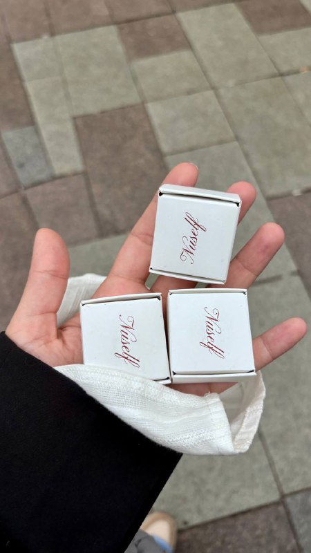 Рука держит маленькие коробочки конфет с логотипом Nuself — три аккуратные коробочки птичьего молока, видна текстура упаковки и рука на фоне улицы.