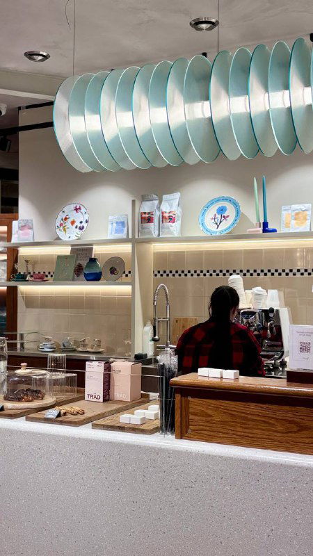 Кофейный прилавок и витрина с тарелками и декором: бариста за стойкой, стильный интерьер с пастельными акцентами и открытой зоной приготовления напитков.