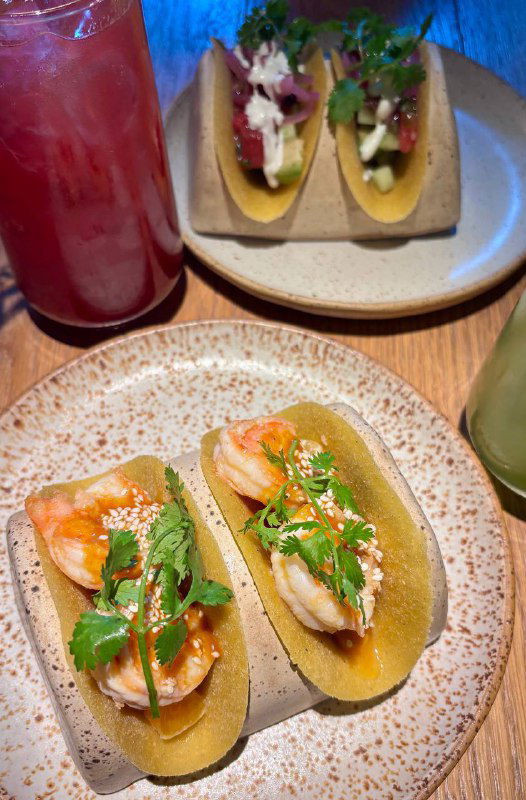 Два тако с креветками и яркими добавками рядом с напитками; аккуратная подача в стиле Tacobar на деревянном столе