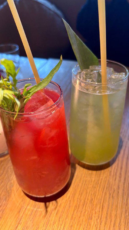 Два цветных коктейля в высоких стаканах с трубочками и зеленью, кадр со столика ресторана Tacobar и тёплым светом