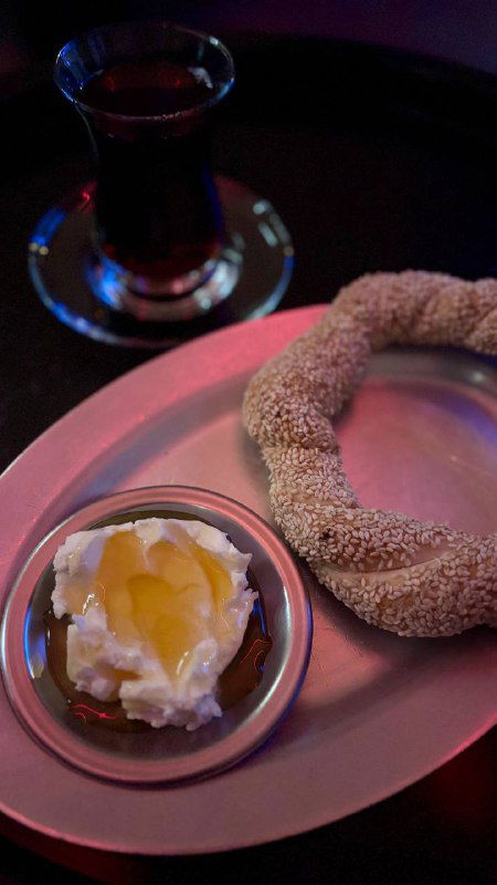 Чай и симит на подносе: бублик в плотной кунжутной корочке, рядом мисочка с густым каймаком и медом, тёплая вечерняя подача.