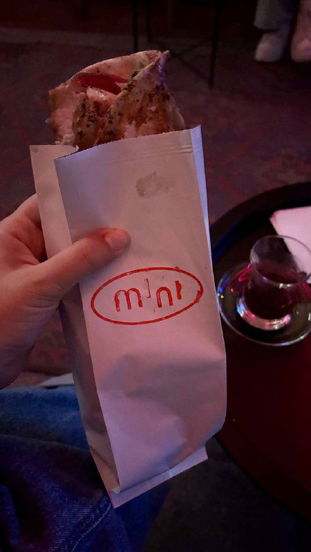 Завёрнутая шаурма/балык в фирменном пакете Mini, на переднем плане рука держит еду, рядом стакан турецкого чая в традиционной форме.