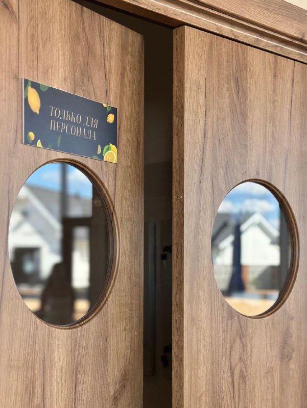 Крупный план двухстворчатых маятниковых дверей с круглыми иллюминаторами: видна фурнитура, зеркало в окне отражает фасады зданий и дневной свет в помещении кафе.