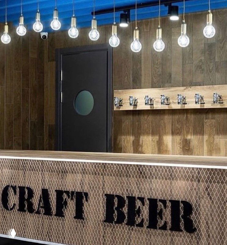 Тёмная маятниковая дверь с круглым иллюминатором за барной стойкой: интерьер в стиле craft beer, деревянные панели на стене и декоративное освещение над стойкой.