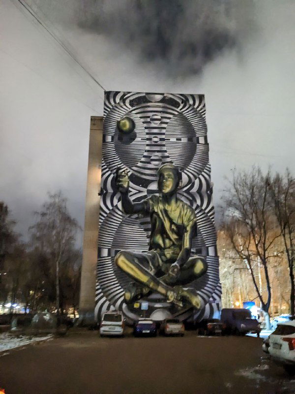 Высокий уличный мурал с оптическими полосами и зелёной сидящей фигурой на переднем плане — контрастный графический приём в вечернем освещении.