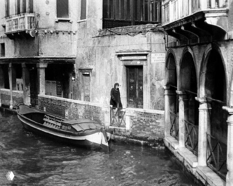 Мужчина идёт по узкой венецианской набережной рядом с гондолой, архитектура старых домов и влажные кирпичные фасады создают гнетущую атмосферу.