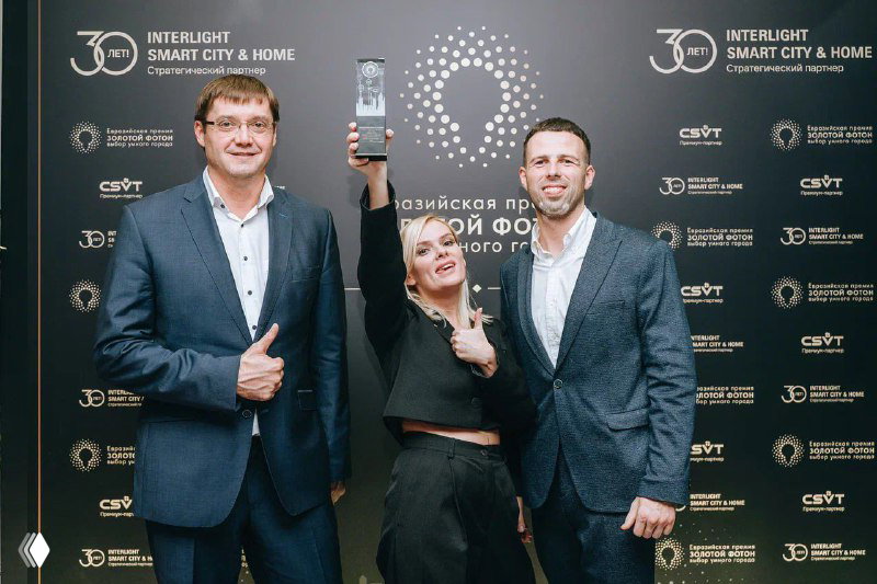 Трое участников держат приз на фоне брендволла Interlight | Smart City & Home — победители позируют с наградой.