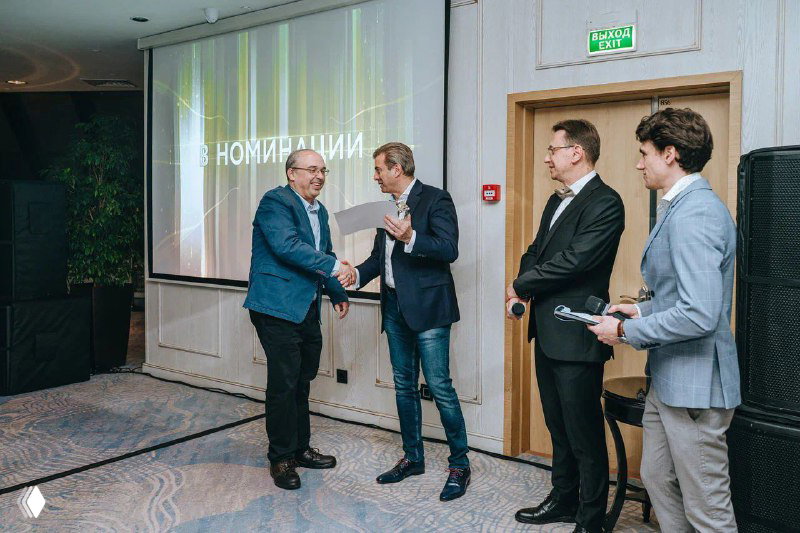 Два человека жмут руки во время церемонии на фоне экрана с номинацией — официальный момент награждения.