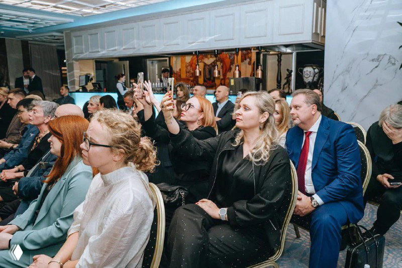 Гости церемонии фотографируют сцену на смартфоны: женщины и мужчины в зале фиксируют моменты награждения.