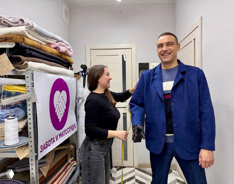 Сотрудница и мужчина примеряют адаптированную одежду в мастерской на фоне стопок тканей и плаката «Забота и милосердие».
