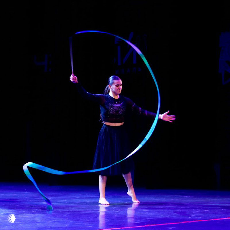 Гимнастка с лентой выполняет сложный элемент на сцене конкурса, крупный план выступления, тёмный фон и акцентное освещение, артистическая поза.