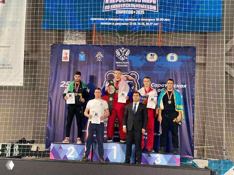 Награждение на пьедестале: спортсмены и тренеры позируют с медалями и кубком перед баннером первенства мира по универсальному бою в Саратове.