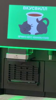 Крупный план экрана с иллюстрацией чашки и подписью «ПРИЯТНОГО КОФЕПИТИЯ», внизу части кофемашины и интерьер офисного пространства.