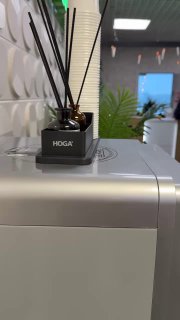 Аромадиффузор HOGA установлен на корпусе кофемашины в офисе: видна маленькая ёмкость с палочками и часть рабочего пространства на заднем плане.