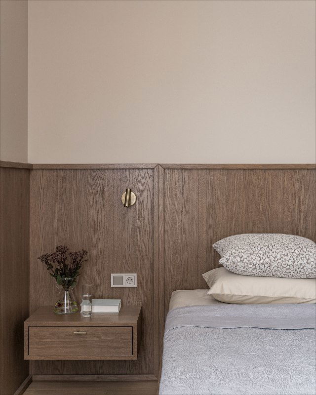 Ближний план прикроватной зоны: изящная деревянная панель, плавающая тумбочка, книги и ваза с сухоцветами — детали, добавляющие уют и текстуру интерьеру.