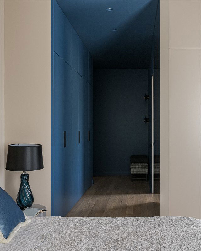 Вид на проход к гардеробной с насыщенными синими фасадами, настольная лампа на тумбе и гармоничная цветовая схема в зоне входа в спальню.