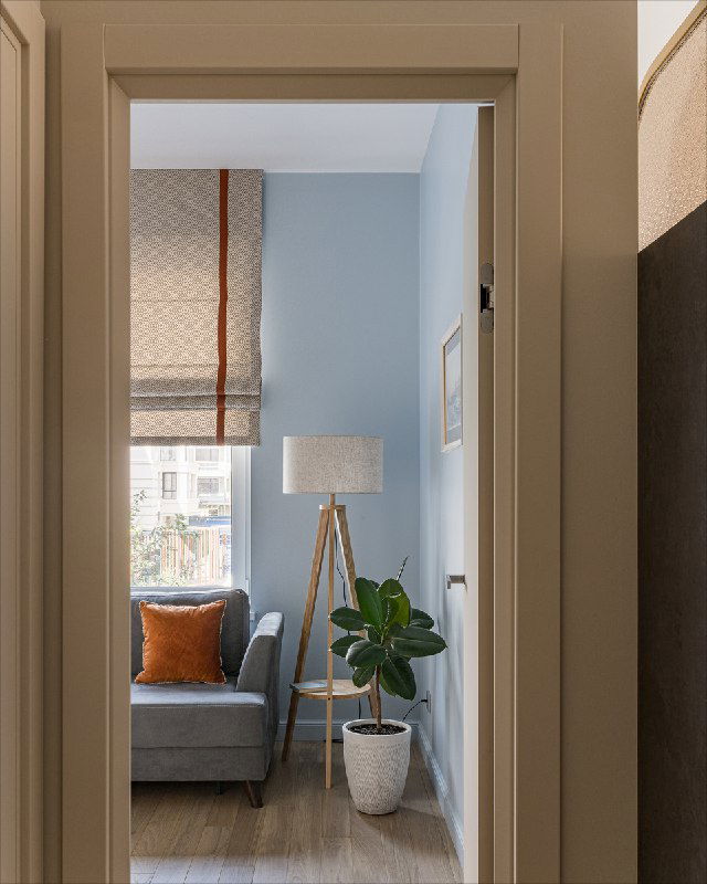 Вид через дверной проем на гостевую комнату: римская штора на окне, диван с оранжевой подушкой, напольный торшер и фикус у нежно-голубой стены.
