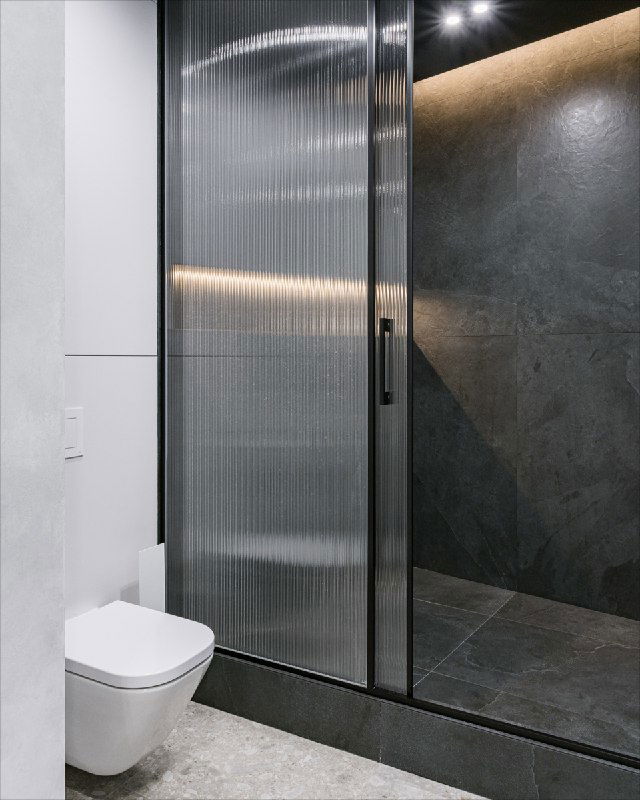 Душевая комната с рифлёным стеклом и тёмной фактурной облицовкой стены; вид на душевую кабину и расположенный рядом санузел.