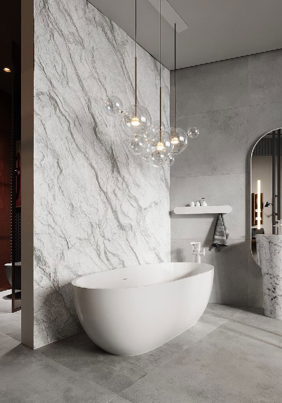 Свободностоящая белая ванна на фоне рельефной мраморной панели, видна стеклянная дверь душевой и воздушная подвесная люстра.